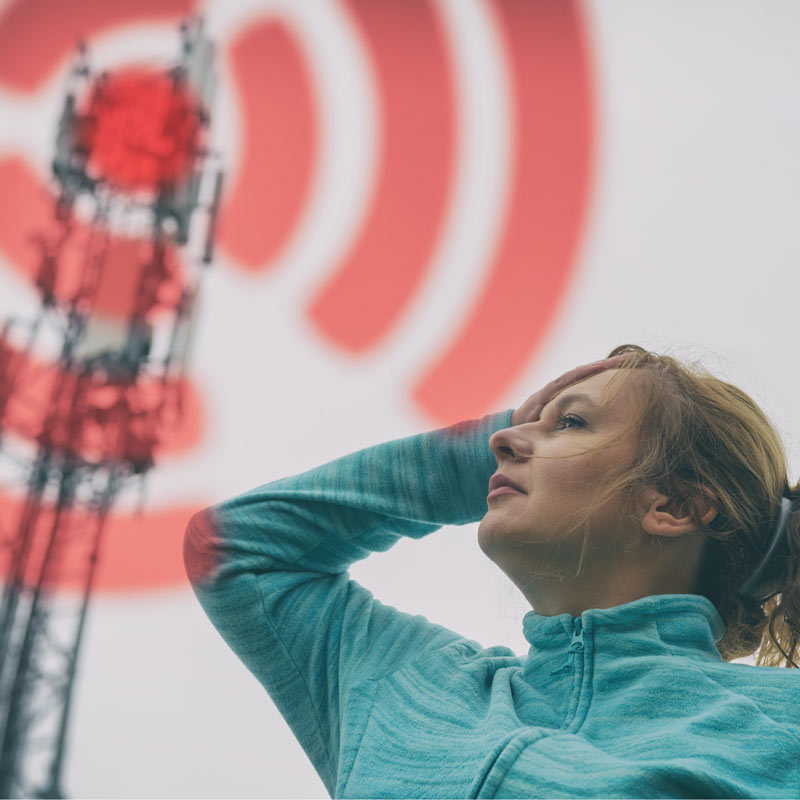 Eine Frau hält vor Schmerzen die Hand an den Kopf vor einer 5G-Antenne